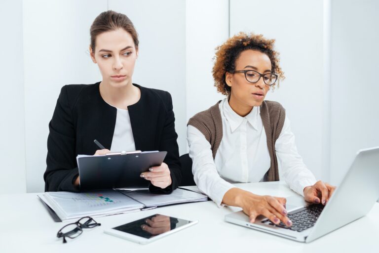 Two focused businesswomen working using clipboard and laptop